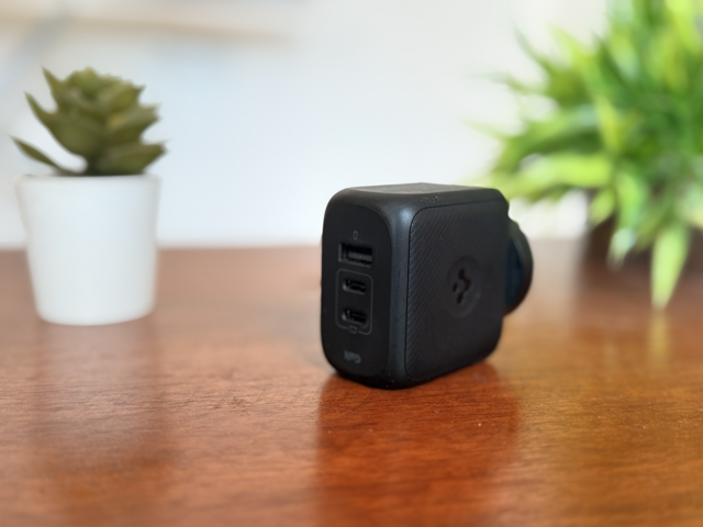 Spigen GaN 65W charger placed on a wooden surface, showcasing its two USB-C ports and one USB-A port, with a blurred background of green plants and a white pot.