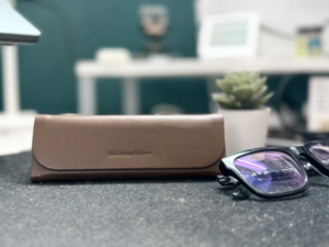 Daily Objects Foldaway Eyewear Case in brown faux leather placed on a textured surface, with a pair of black-framed glasses and a small potted plant in the background, showcasing its sleek and minimalist design.